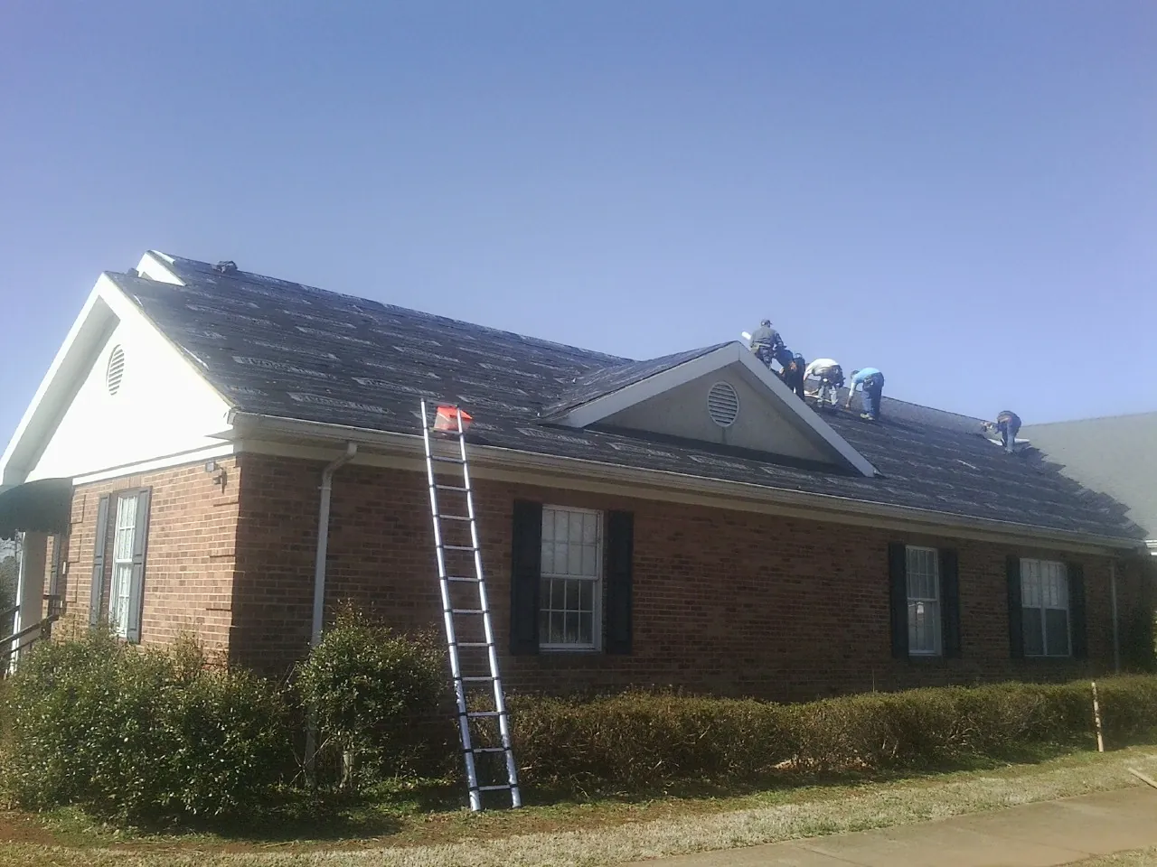 Crew tearing shingles off a brick ranch roof with a ladder set — Emergency Roof Repair work by our crew in Fonda
