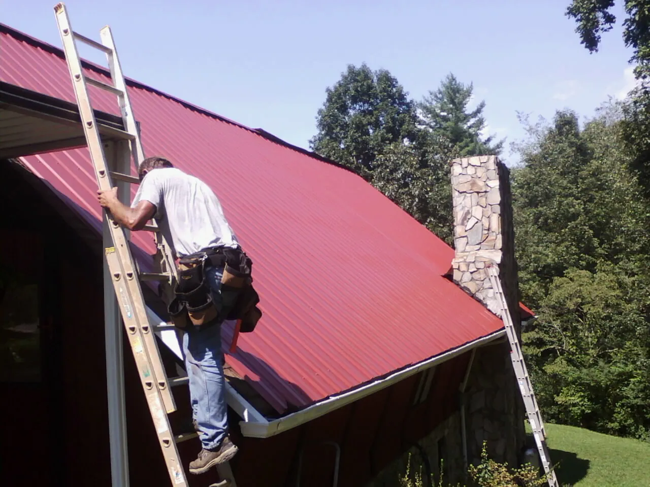 Roofer on a ladder beside a red metal roof and stone chimney — Soffit & Fascia Repair by Roofing Brothers in Fonda